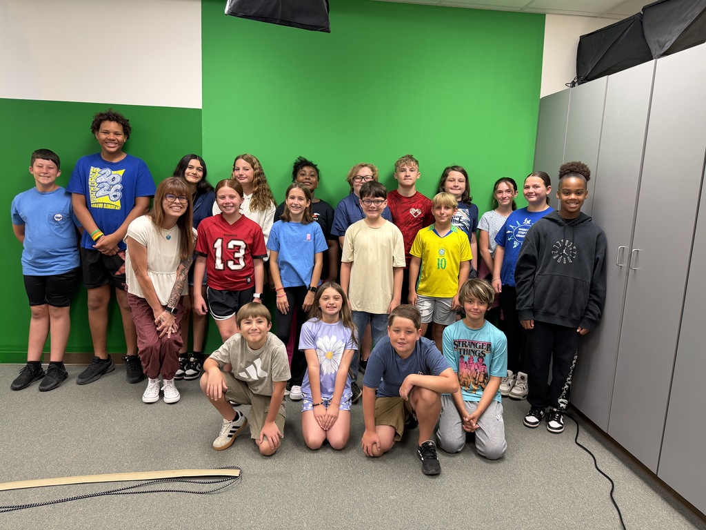 A group photo of diverse children posing in front of a large green screen. An adult woman with glasses and tattoos smiles and kneels on the left side of the front row alongside four kneeling children, with the rest of the group standing behind them.