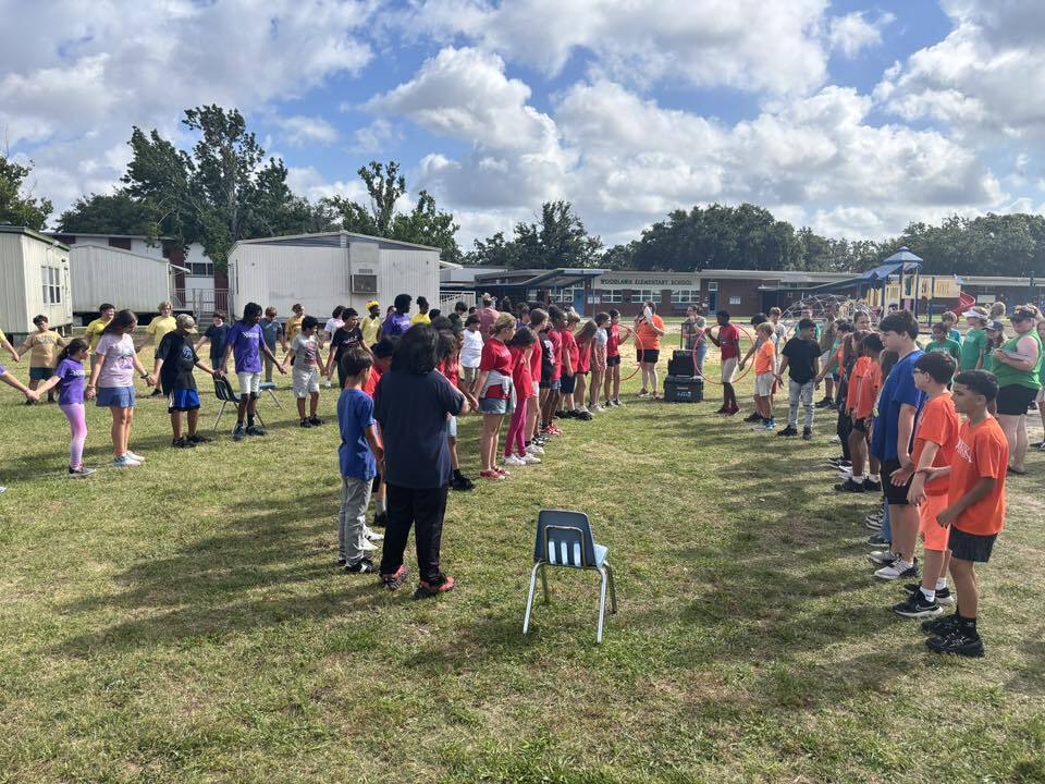 A large group of diverse elementary school students stand in a wide circle holding hands on a grassy field, participating in an outdoor activity near school buildings.