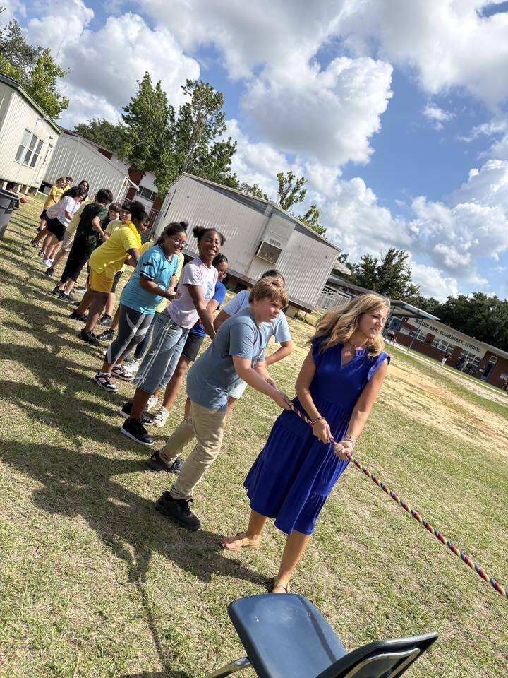 A woman in a blue dress and a line of elementary students pull a colorful rope during a tug-of-war game on a grassy school field under a bright, cloudy sky.