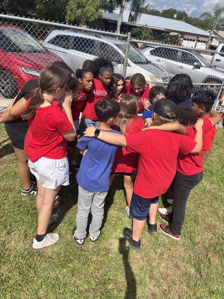 A tight huddle of elementary school children wearing red and blue shirts put their arms around each other on a grassy field near a chain-link fence.