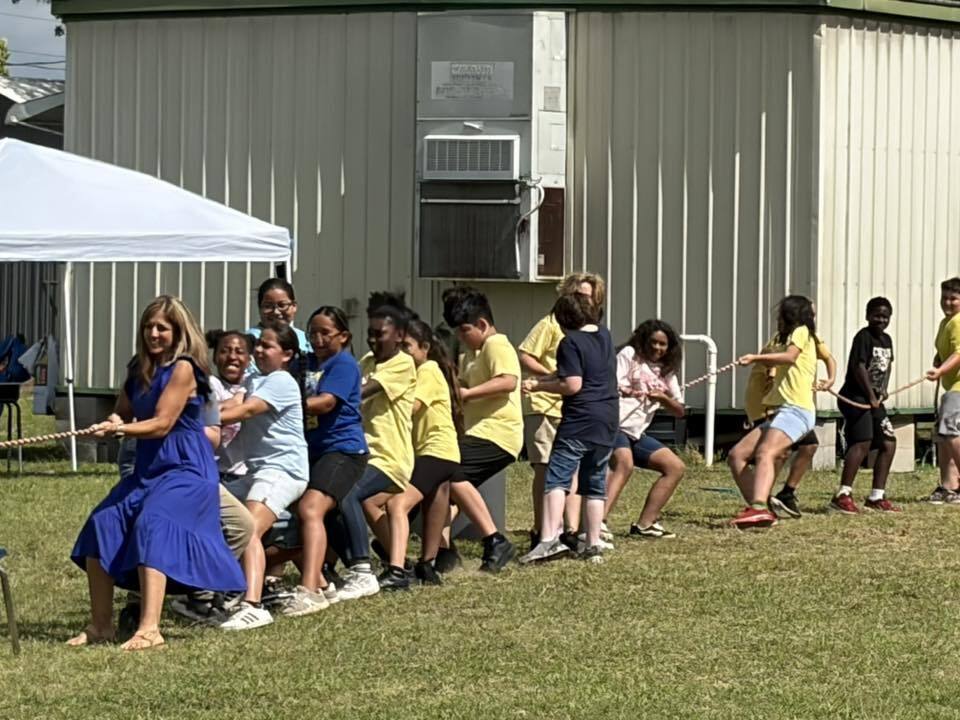 A woman in a blue dress and a line of children in yellow, blue, and black shirts strain together while pulling a rope during an outdoor tug-of-war match.