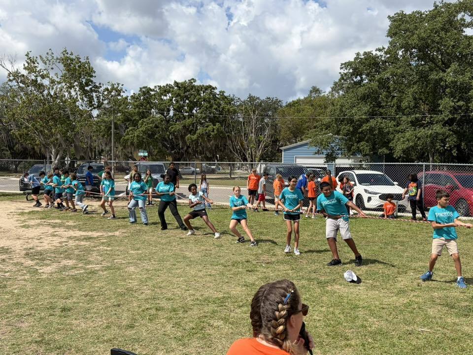 A team of elementary school children wearing matching teal shirts pull hard on a rope during a tug-of-war competition on a grassy field lined with parked cars.