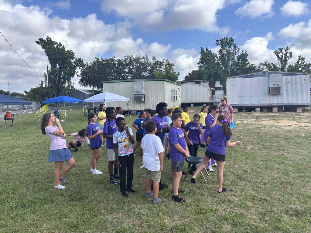 A group of children, many wearing matching purple shirts, gather and talk on a grassy field during an outdoor school event with portable classrooms in the background.