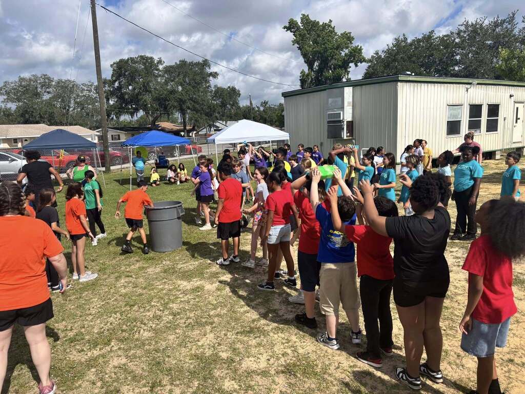 A line of elementary students plays an outdoor water game, passing objects over their heads down a queue toward a large gray plastic bin on a grassy field.