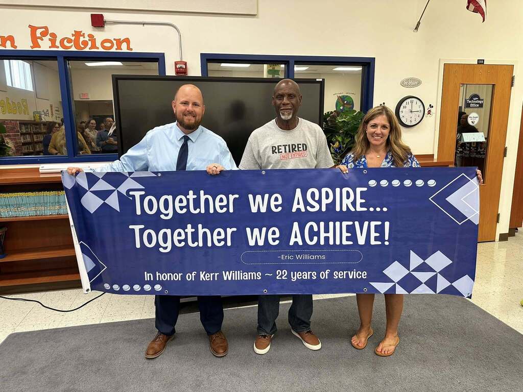 Three people stand in a school media center holding a large blue banner that reads, "Together we ASPIRE... Together we ACHIEVE! –Eric Williams. In honor of Kerr Williams ~ 22 years of service." The center man wears a grey t-shirt that reads, "RETIRED NOT EXPIRED."