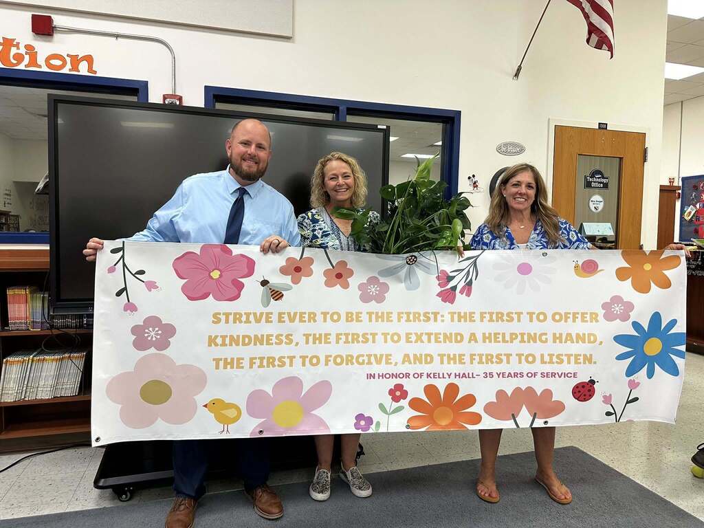 Three people stand in a school media center holding a large white banner decorated with colorful flowers, a bee, and a ladybug. The banner reads, "STRIVE EVER TO BE THE FIRST... IN HONOR OF KELLY HALL– 35 YEARS OF SERVICE."