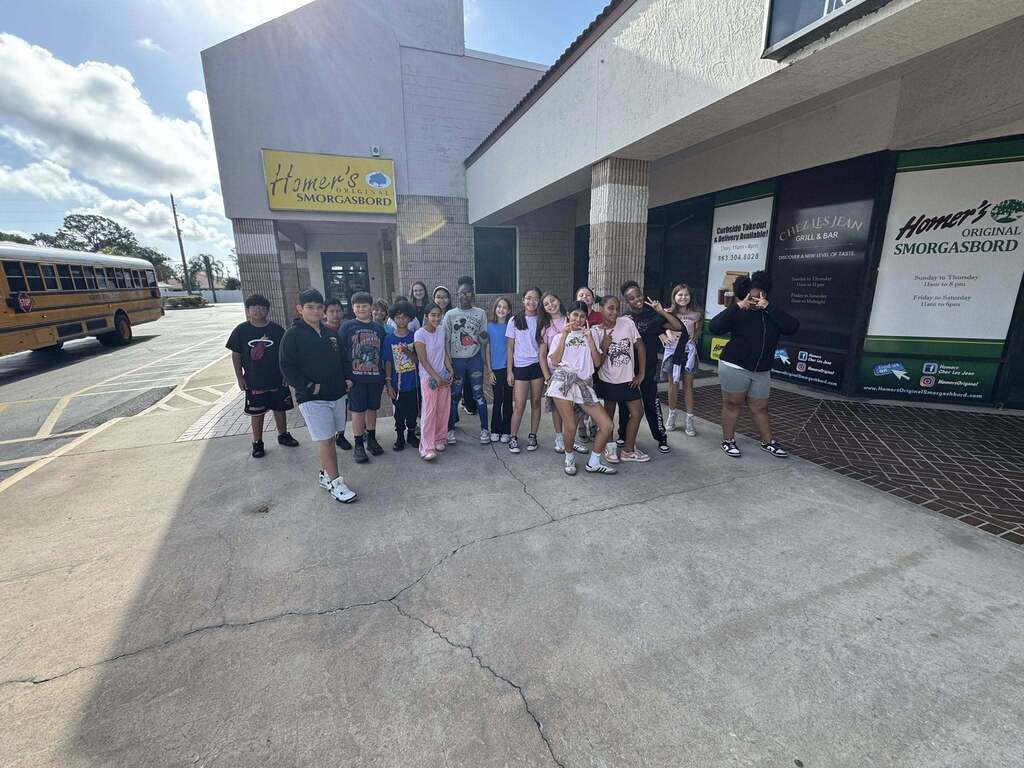 A wide-angle, eye-level outdoor shot shows a large group of young students standing together on a concrete walkway in front of "Homer’s Original Smorgasbord." A yellow school bus is parked to the left. The kids are dressed casually, smiling and posing for the camera on a sunny day.