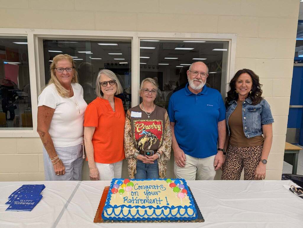 Five people stand side-by-side behind a long table at a retirement celebration. A large rectangular cake sits in the center with blue frosting borders and text reading, "Congrats on your Retirement!" Blue napkins are on the left. The background features a light-colored wall with large windows.