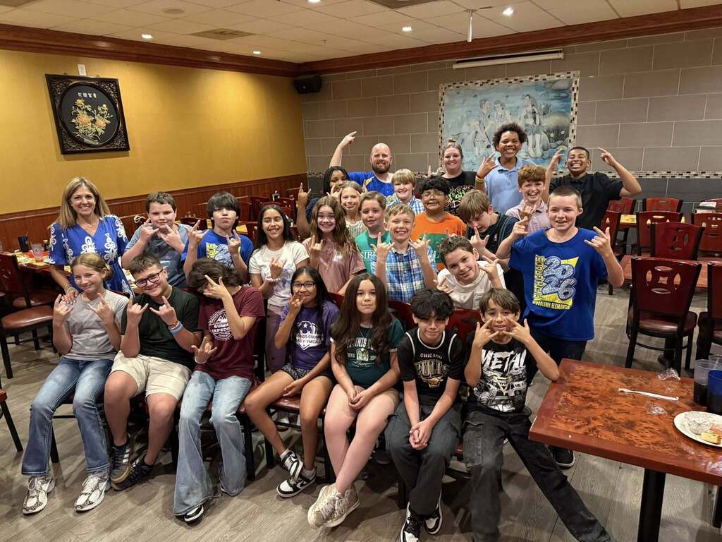 A large group of children and three adults pose together in a restaurant, many smiling and making hand gestures. They are seated and standing in multiple rows in front of a yellow and grey wall. Tables, chairs, and restaurant decor are visible in the background.