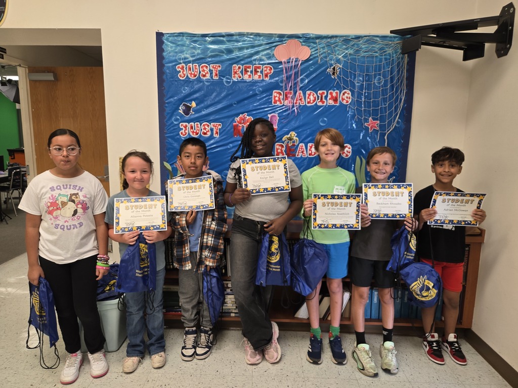 Seven young students stand in a row in a school hallway, posing with "Student of the Month" certificates and blue drawstring bags. Behind them is a low wooden bookcase and a blue bulletin board reading "JUST KEEP READING."