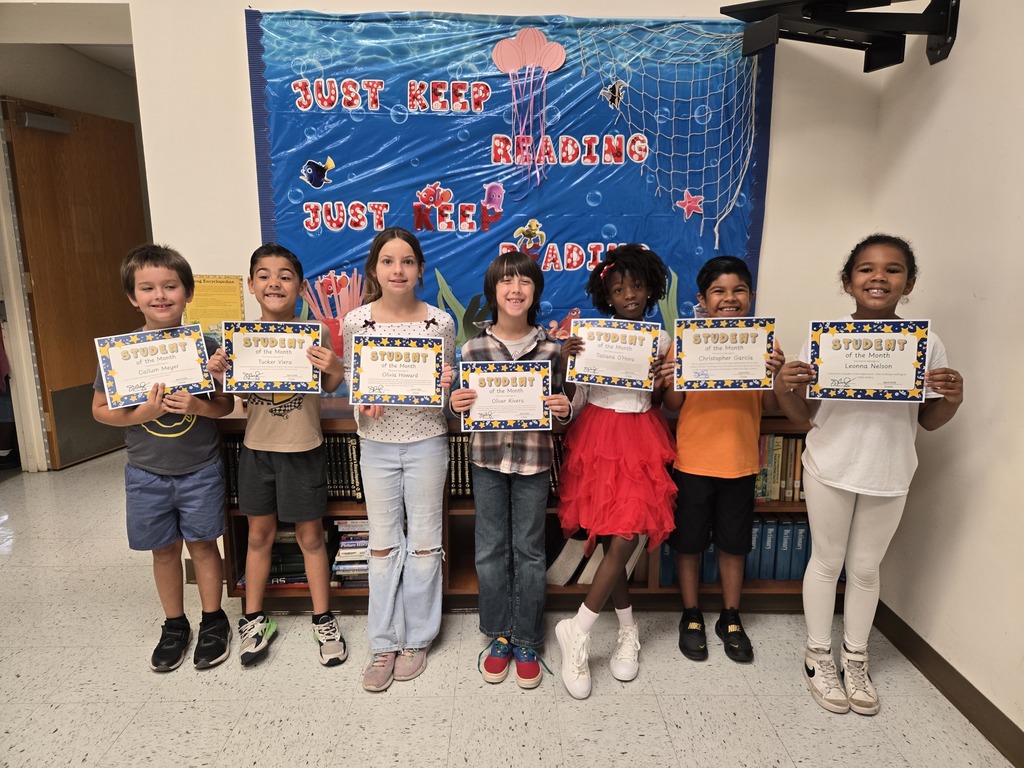 Seven elementary-aged students stand in a line holding "Student of the Month" certificates. They are posing in front of a blue school bulletin board that reads "JUST KEEP READING." A low bookshelf with books is visible behind them.