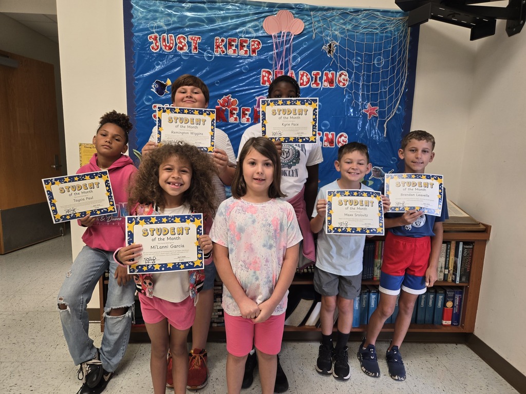 Seven diverse young students stand close together in two informal rows, proudly holding up their "Student of the Month" certificates. They are posing in front of a blue "JUST KEEP READING" themed bulletin board and a bookshelf.