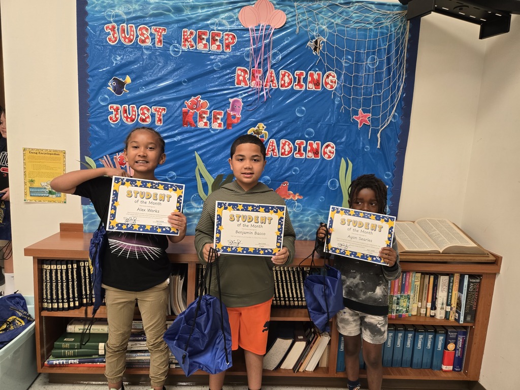 Three elementary-aged students stand side-by-side in front of a blue "JUST KEEP READING" bulletin board. Each child holds a "Student of the Month" certificate and a blue drawstring backpack while smiling at the camera.