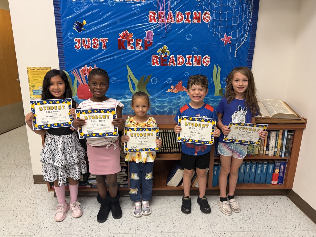 Five elementary-aged students stand in a line in front of a blue ocean-themed bulletin board that says "JUST KEEP READING." Each smiling student is holding up a "Student of the Month" certificate for the camera.