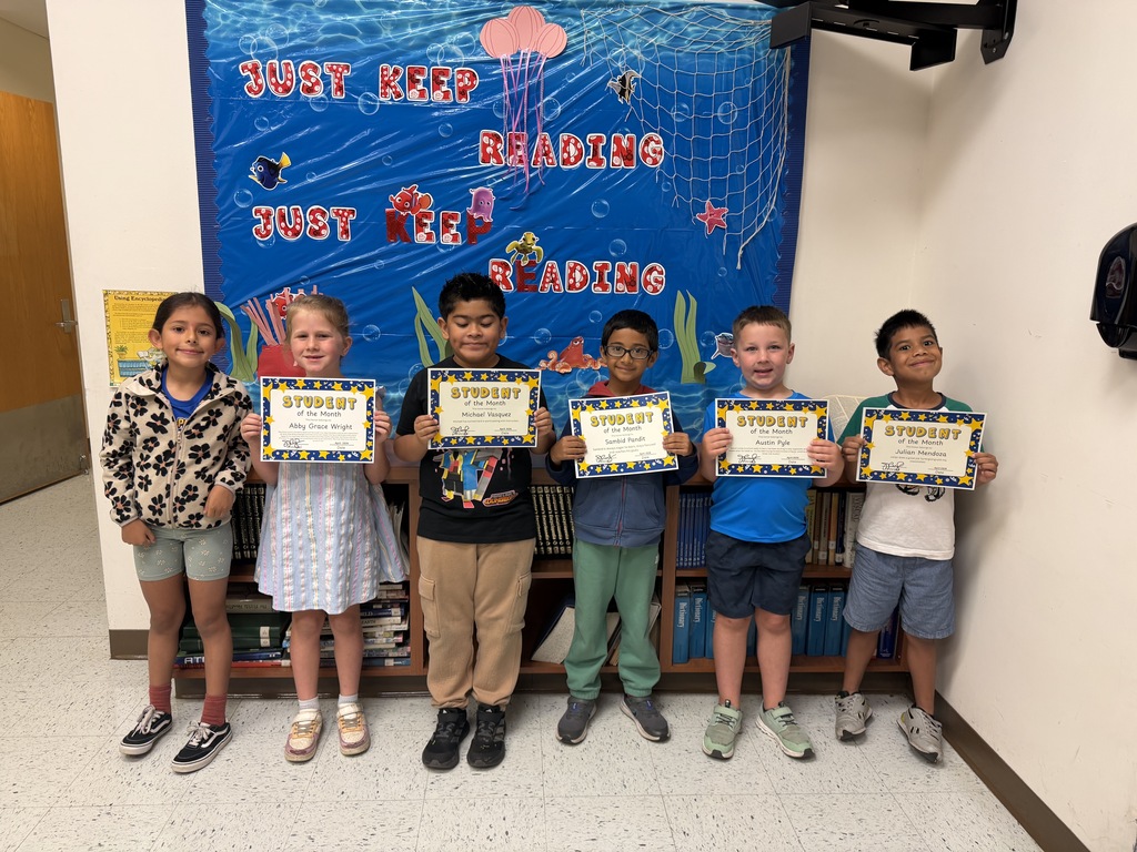 Six young students stand side-by-side in a school hallway, posing proudly. Five of the children hold up "Student of the Month" certificates. Behind them is a wooden bookcase and a blue "JUST KEEP READING" bulletin board.