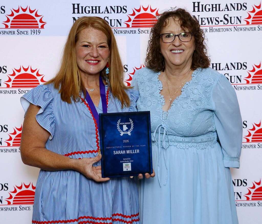 Two smiling women stand together holding a plaque that reads "Highlander Award 2026 Agricultural Person of the Year SARAH MILLER." The woman on the left wears a blue-and-white striped dress, and the woman on the right wears a light blue lace dress and glasses, standing before a branded backdrop.