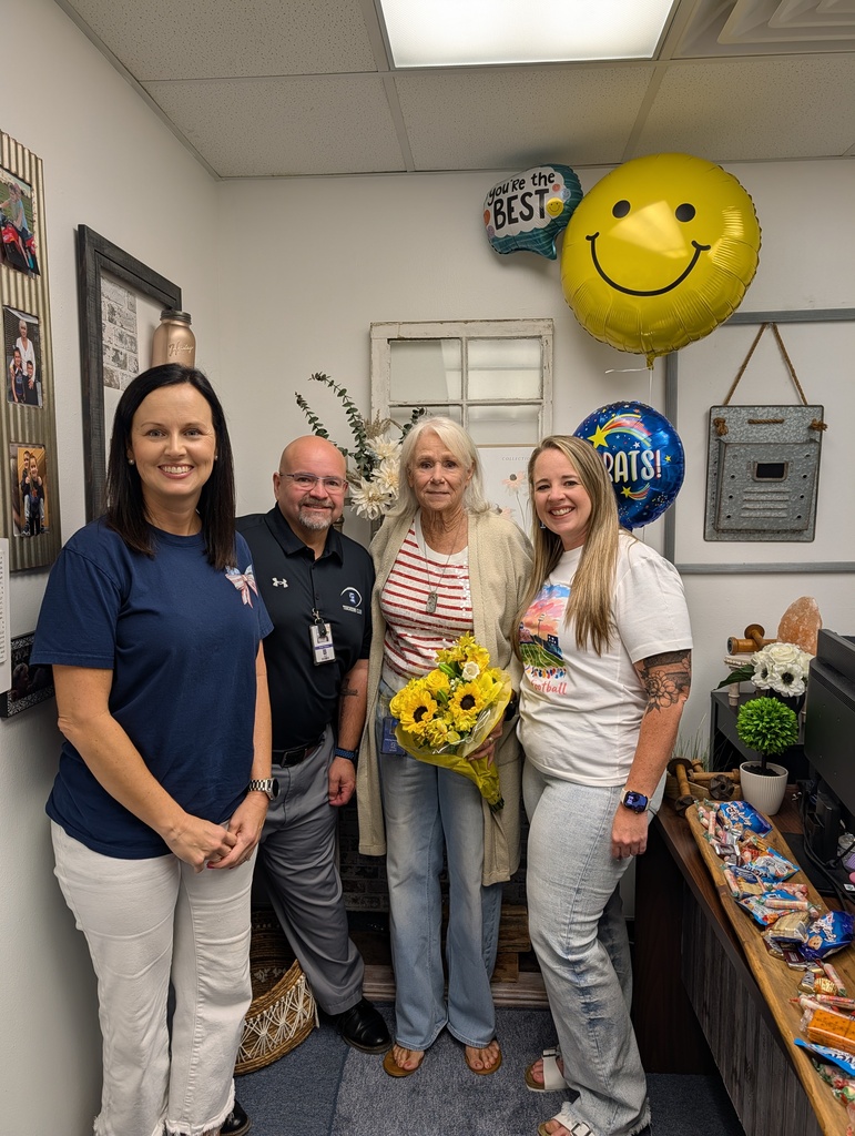 Four colleagues pose smiling inside an office space. A central woman with blonde hair holds a bouquet of yellow sunflowers while standing alongside three coworkers. Above them float celebratory balloons, including a large yellow smiley face, "Congrats!", and "You're the BEST" balloons.