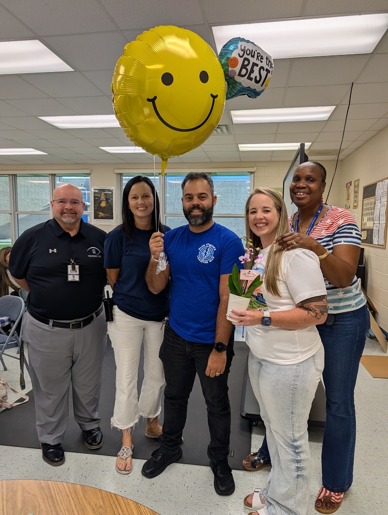 Five smiling adults stand together indoors in a brightly lit room. The man in the center holds strings attached to a large yellow smiley-face balloon and a "You're the Best" foil balloon. The woman beside him holds a potted orchid, while three colleagues pose around them in celebration.