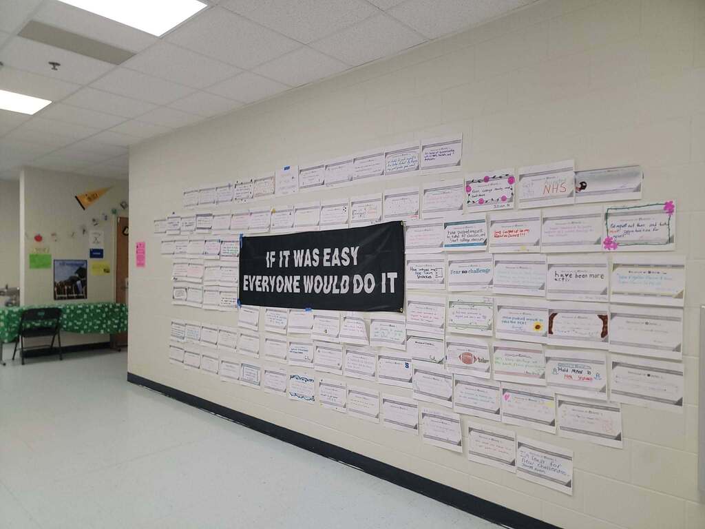 A classroom hallway wall covered in dozens of small paper notes with handwritten messages. A prominent black banner hung in the center reads, "IF IT WAS EASY EVERYONE WOULD DO IT" in white capital letters.