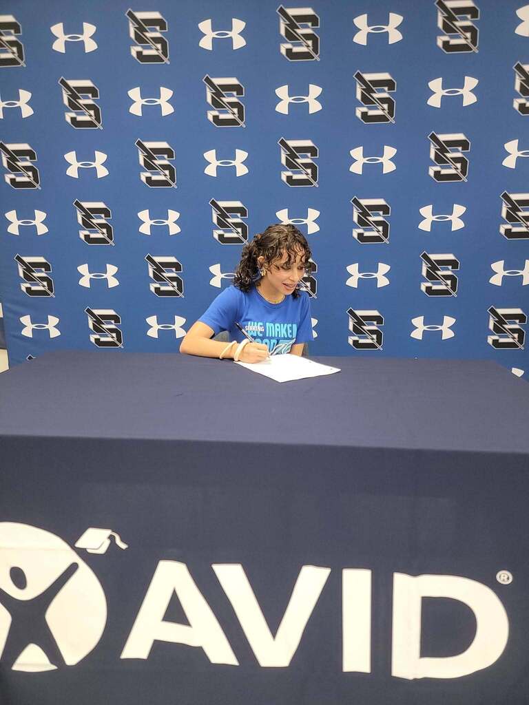 A young woman with curly hair wearing a blue t-shirt sits at a table covered in a dark blue tablecloth with a prominent white AVID logo. She holds a pen and fills out a paper on the table in front of a blue backdrop.