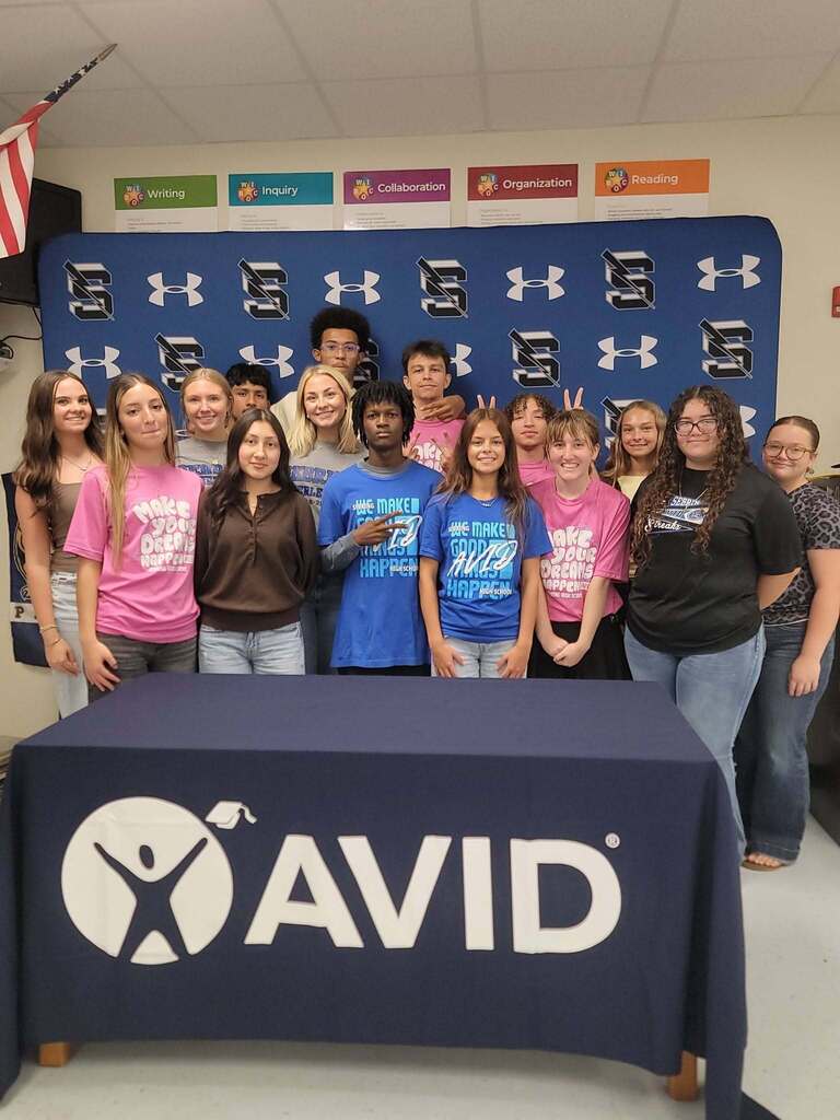 A group of high school students stands behind a table draped with a dark blue tablecloth featuring a large white AVID logo. The students are dressed in casual attire, with several wearing pink and blue AVID t-shirts, posing in front of a branded blue backdrop.