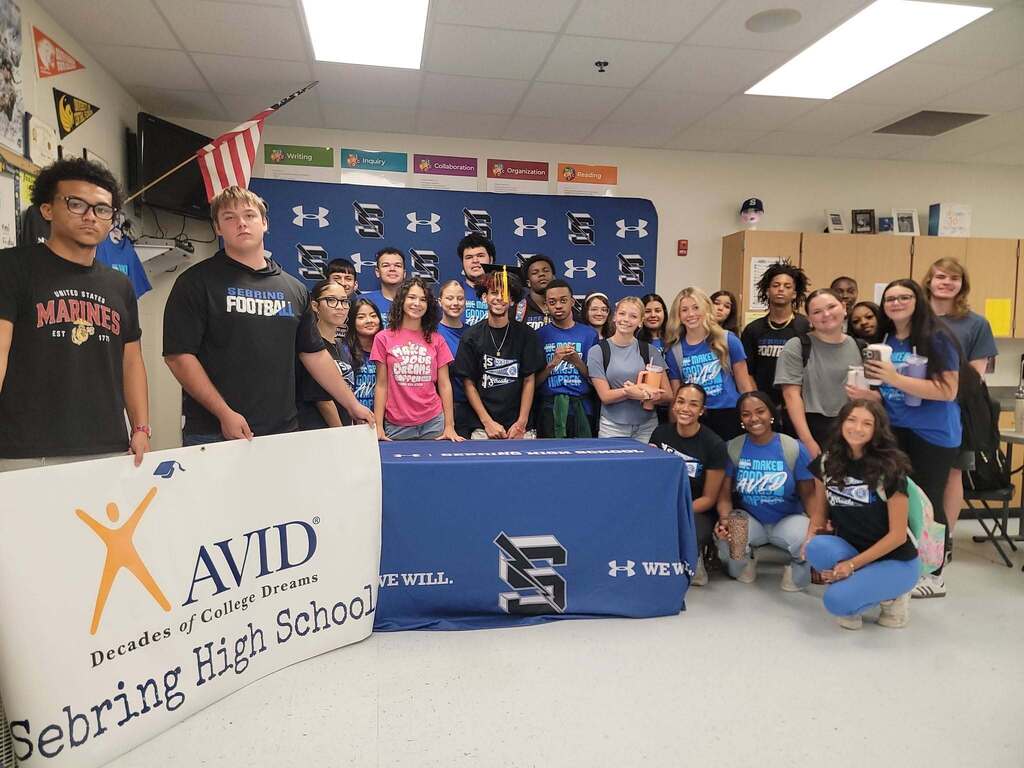 A large group of high school students poses together in a classroom. In the foreground on the left, two students hold a white banner reading "AVID Decades of College Dreams - Sebring High School." Behind them, students stand in rows in front of a blue Under Armour backdrop.