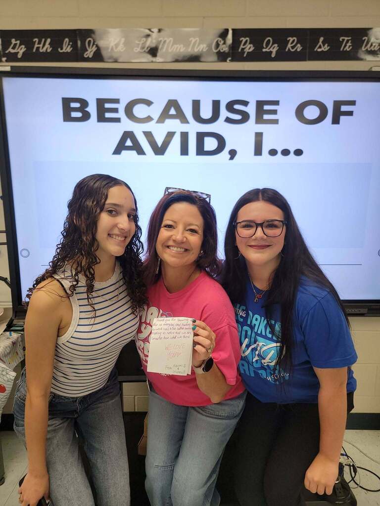 Three women stand close together in front of a digital screen displaying "BECAUSE OF AVID, I...". The middle woman wears a bright pink shirt and holds a handwritten thank-you card. The woman on the left wears a striped sleeveless top, and the woman on the right wears a blue AVID t-shirt and glasses.