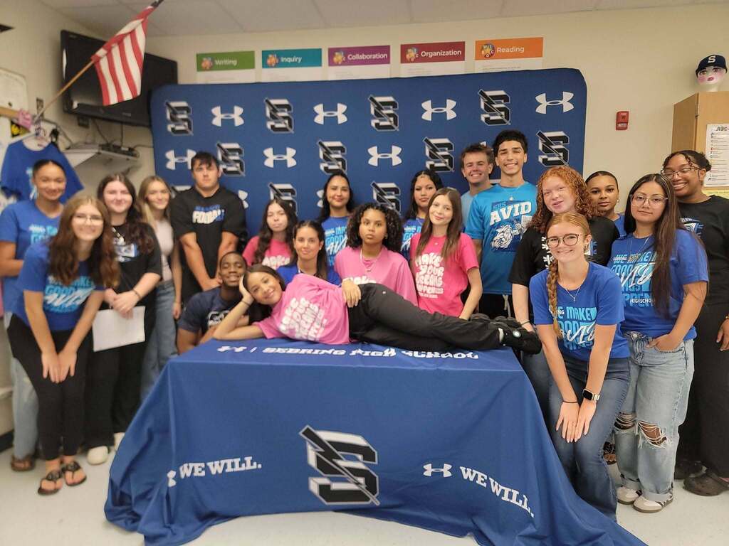A group of high school students poses around a table covered with a blue "Sebring High School" cloth featuring an AVID logo. One student in pink lies across the top of the table posing, while other students stand and sit around her, many wearing blue or pink matching t-shirts.