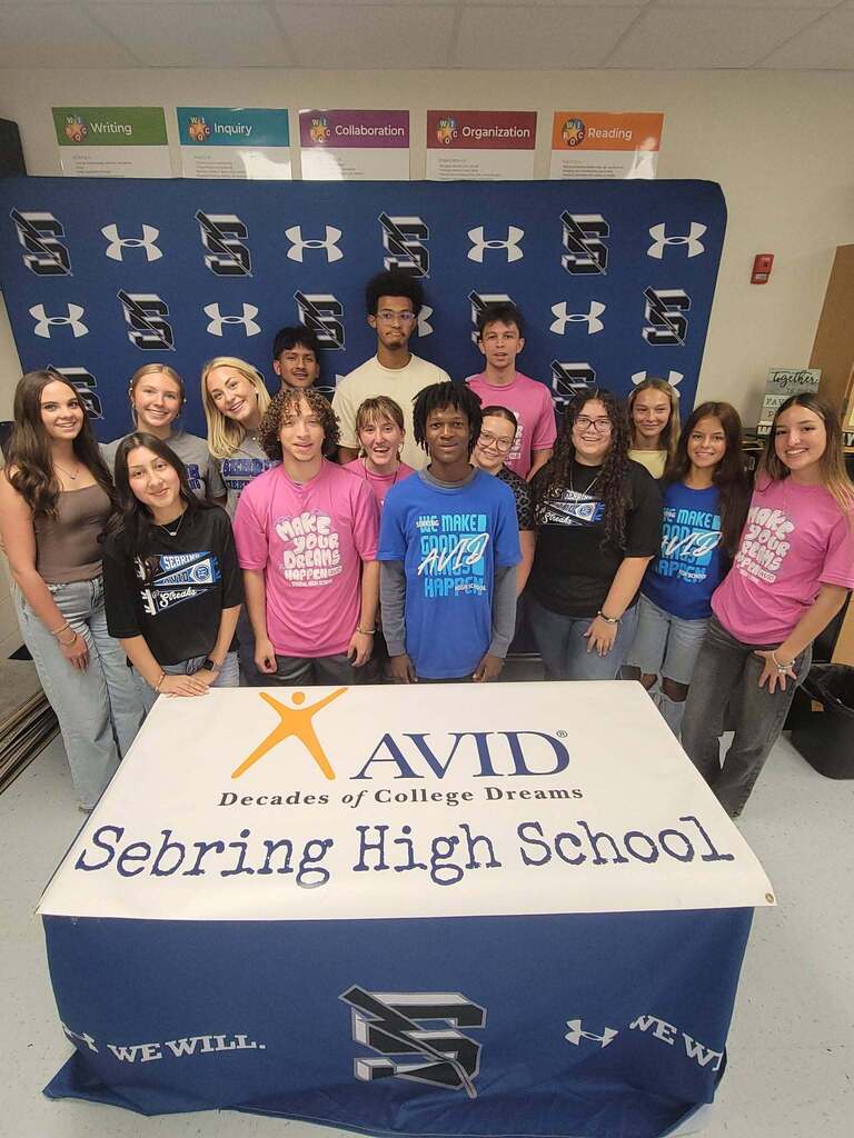 A group of 14 students poses standing in rows behind a table draped with a blue and white "AVID - Sebring High School" runner. They stand in front of a blue backdrop featuring repeating Under Armour logos and educational posters reading "Writing," "Inquiry," "Collaboration," "Organization," and "Reading."