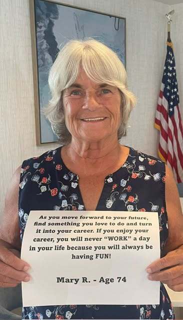 A smiling woman with blonde hair wearing a patterned top holds a sign reading: "As you move forward to your future, find something you love to do and turn it into your career. If you enjoy your career, you will never 'WORK' a day in your life because you will always be having FUN! Mary R. - Age 74."