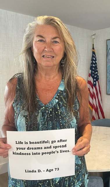 A woman with long blonde hair wearing a blue patterned sleeveless top holds a sign reading: "Life is beautiful; go after your dreams and spread kindness into people’s lives. Linda D. - Age 73." An American flag stands in the background.