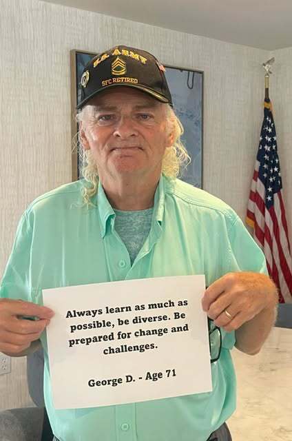 A man wearing a black U.S. Army SFC Retired cap and a mint green shirt holds a sign reading: "Always learn as much as possible, be diverse. Be prepared for change and challenges. George D. - Age 71." An American flag stands in the background.