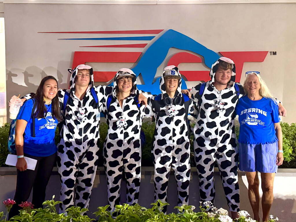 Four swimmers in cow onesies stand between two coaches in blue team shirts beneath the FAST logo.