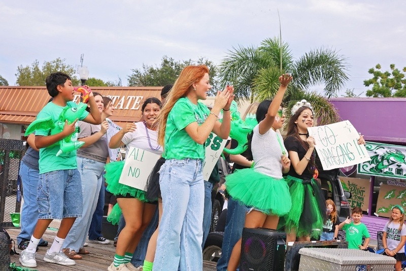 Students in green tutus cheering on a parade float with inflatable dragons and signs.