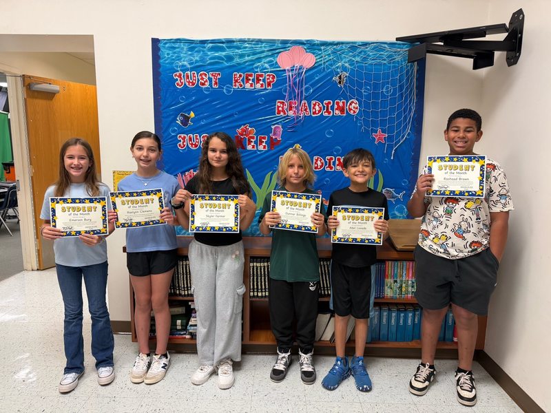 young children stand in a hallway in front of an underwater-themed "Just Keep Reading" display, each proudly holding a "Student of the Month" certificate.