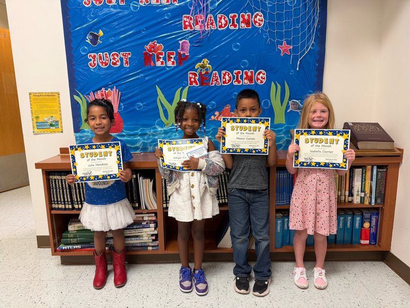 young children stand in a hallway in front of an underwater-themed "Just Keep Reading" display, each proudly holding a "Student of the Month" certificate.