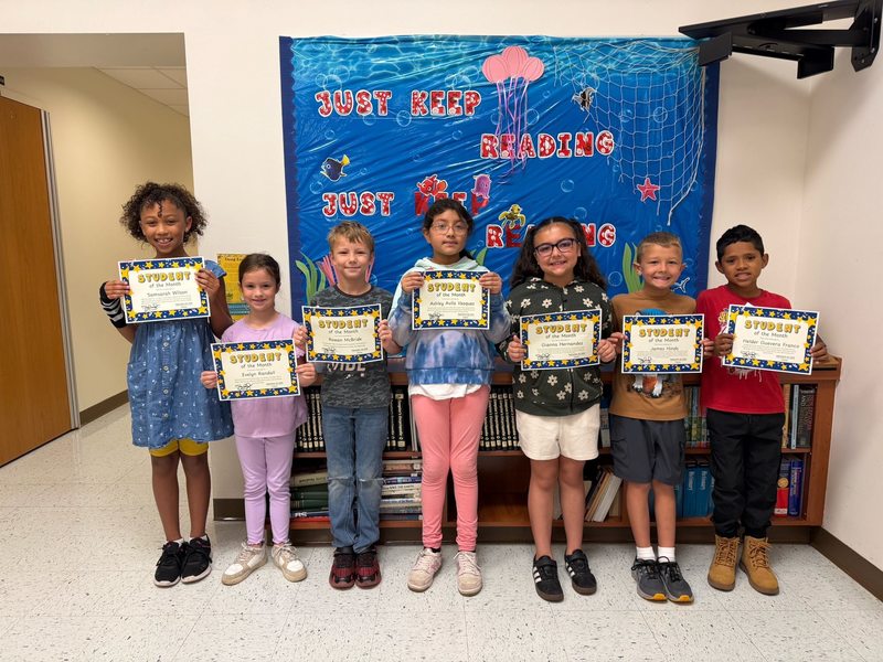 young children stand in a hallway in front of an underwater-themed "Just Keep Reading" display, each proudly holding a "Student of the Month" certificate.
