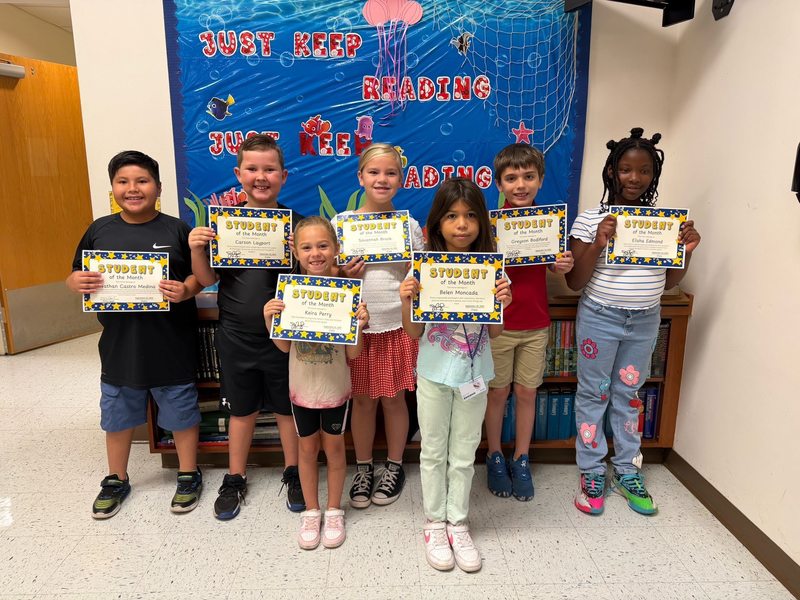 young children stand in a hallway in front of an underwater-themed "Just Keep Reading" display, each proudly holding a "Student of the Month" certificate.