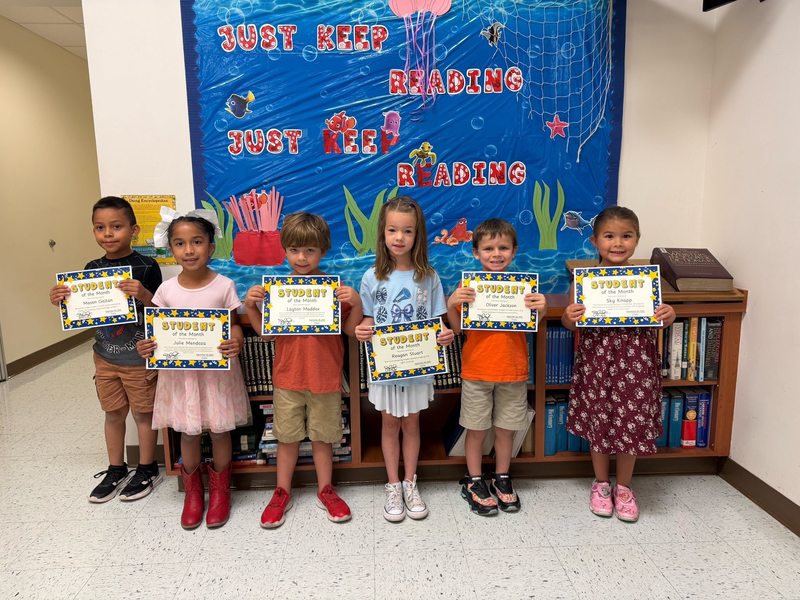 young children stand in a hallway in front of an underwater-themed "Just Keep Reading" display, each proudly holding a "Student of the Month" certificate.