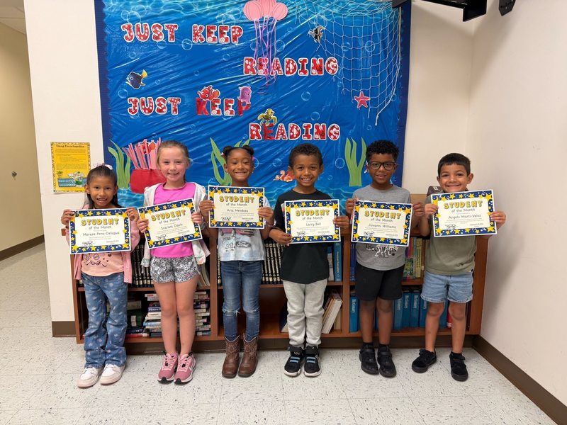 young children stand in a hallway in front of an underwater-themed "Just Keep Reading" display, each proudly holding a "Student of the Month" certificate.
