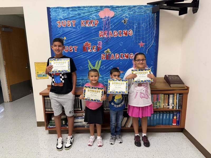 young children stand in a hallway in front of an underwater-themed "Just Keep Reading" display, each proudly holding a "Student of the Month" certificate.