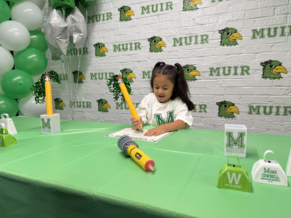 A student holding a giant pecil writing their name on a certificate.