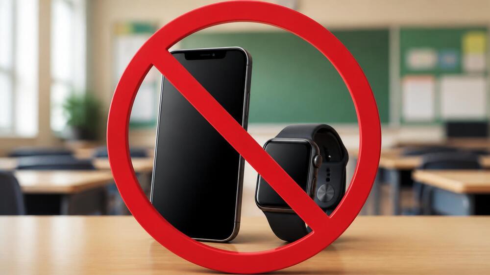 A smart phone and smart watch are sitting upright on top of a desk in a classroom.  There is a red no symbol over the devices.