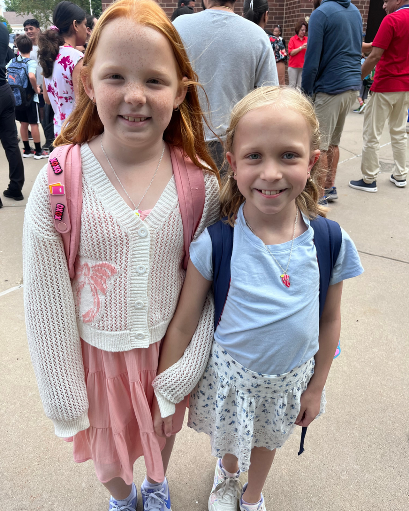 2 student girls smiling together outside with backpacks on before their first day