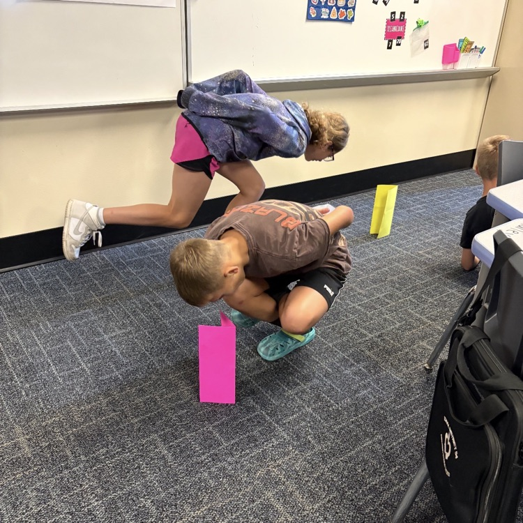 Two 6th graders balancing trying to pick up a paper.