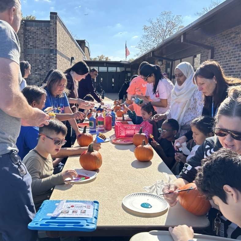 students, teachers, and parents sitting around a table painting pumpkins 