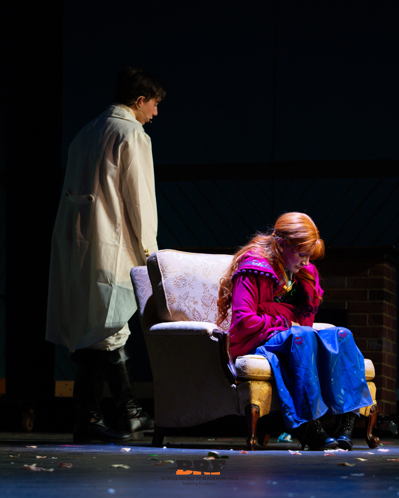 Two actors on the stage, on sitting and the other standing up and looking down.