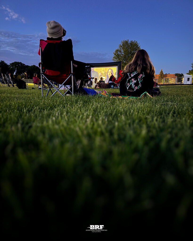 Students sit on the football field, watching a movie.