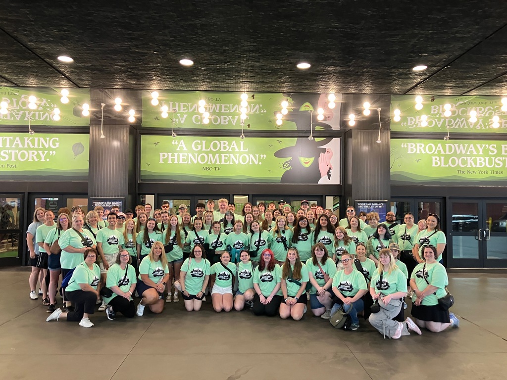 Large group of people wearing green t-shirts pose in front of a sign for Wicked.