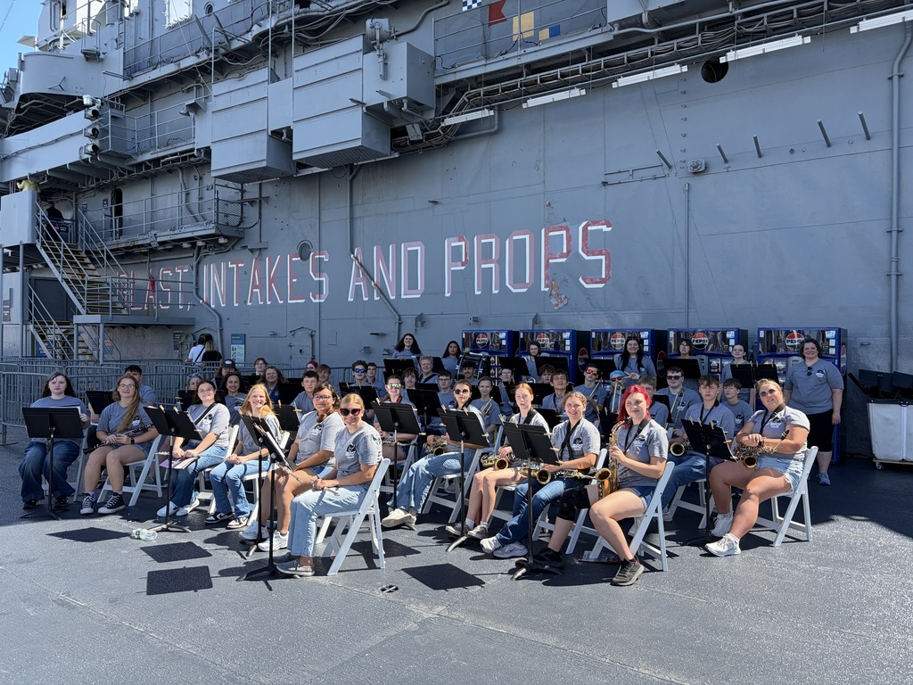 Students holding instruments and sitting on chairs in front of a ship look at camera 